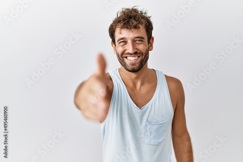 Young handsome man standing over isolated background smiling friendly offering handshake as greeting and welcoming. successful business.