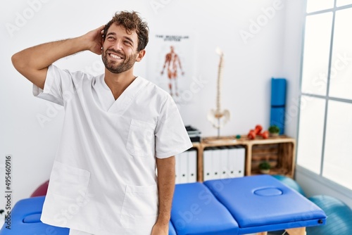 Young handsome physiotherapist man working at pain recovery clinic smiling confident touching hair with hand up gesture  posing attractive and fashionable