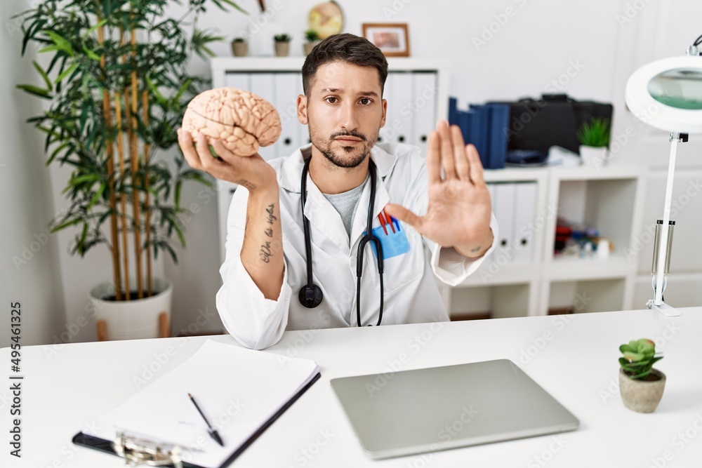 Young doctor holding brain at medical clinic with open hand doing stop sign with serious and confident expression, defense gesture