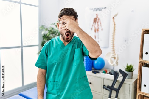 Young man with beard working at pain recovery clinic peeking in shock covering face and eyes with hand, looking through fingers with embarrassed expression.