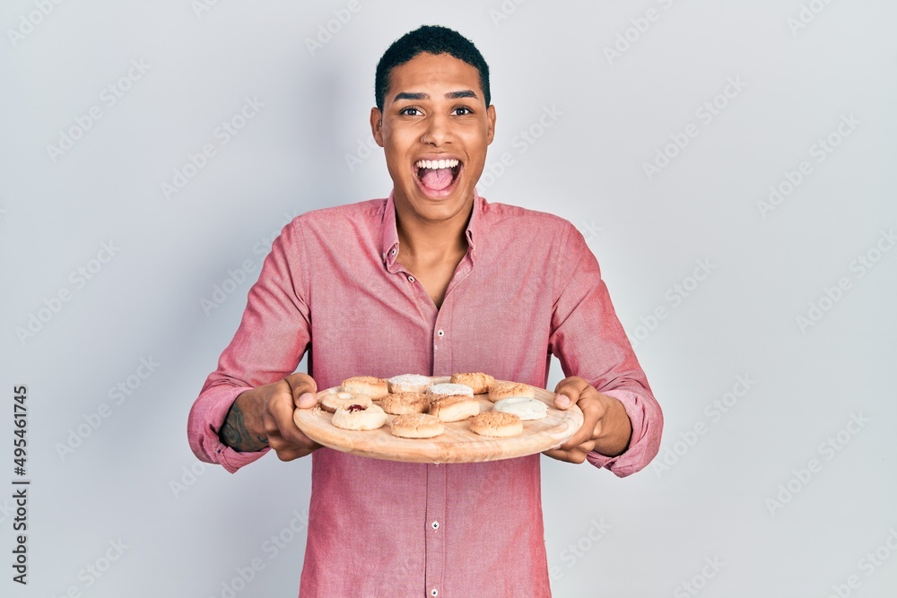 Young african american guy holding tray with cake sweets celebrating crazy and amazed for success with open eyes screaming excited. Young african american guy holding tray with cake sweets celebrating crazy and amazed for success with open eyes screaming excited.
