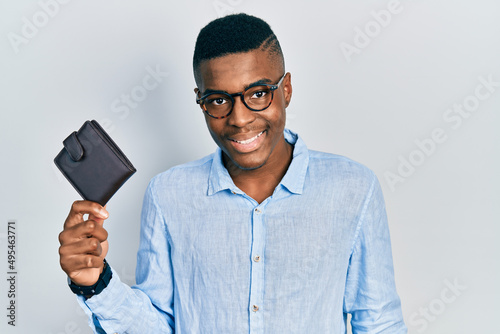 Young african american man wearing glasses holding leather wallet looking positive and happy standing and smiling with a confident smile showing teeth © Krakenimages.com