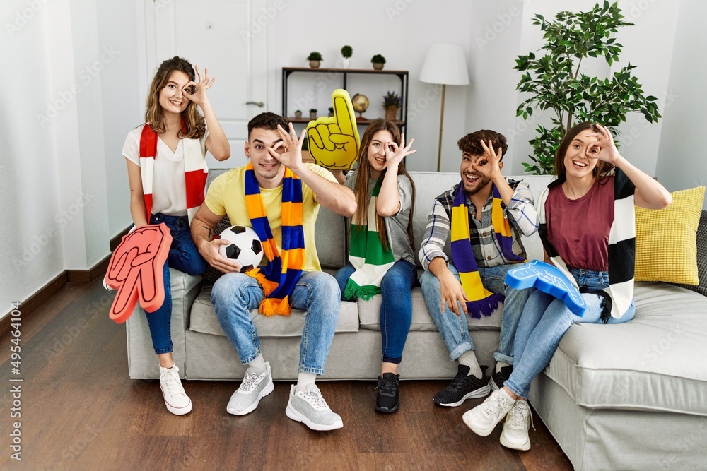 Fototapeta premium Group of young people watching football game wearing team scarf cheering game sitting on the sofa smiling happy doing ok sign with hand on eye looking through fingers