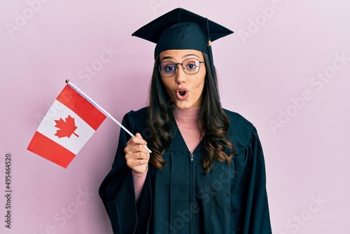 Young hispanic woman wearing graduation uniform holding canada flag scared and amazed with open mouth for surprise, disbelief face