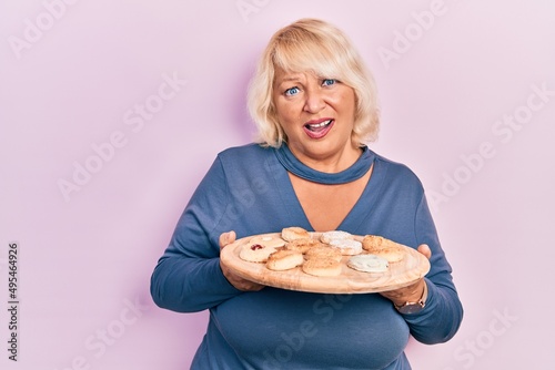Middle age blonde woman holding tray with cake sweets in shock face  looking skeptical and sarcastic  surprised with open mouth