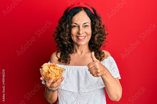 Middle age hispanic woman holding potato chip smiling happy and positive  thumb up doing excellent and approval sign