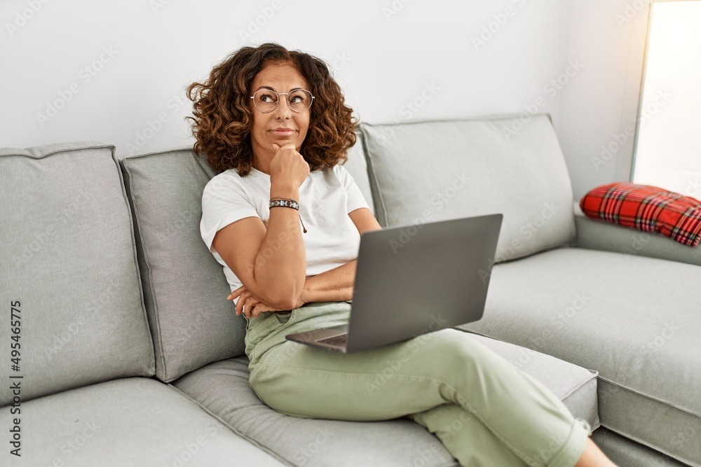 Middle age hispanic woman working using computer laptop at home serious face thinking about question with hand on chin, thoughtful about confusing idea Middle age hispanic woman working using computer laptop at home serious face thinking about question with hand on chin, thoughtful about confusing idea