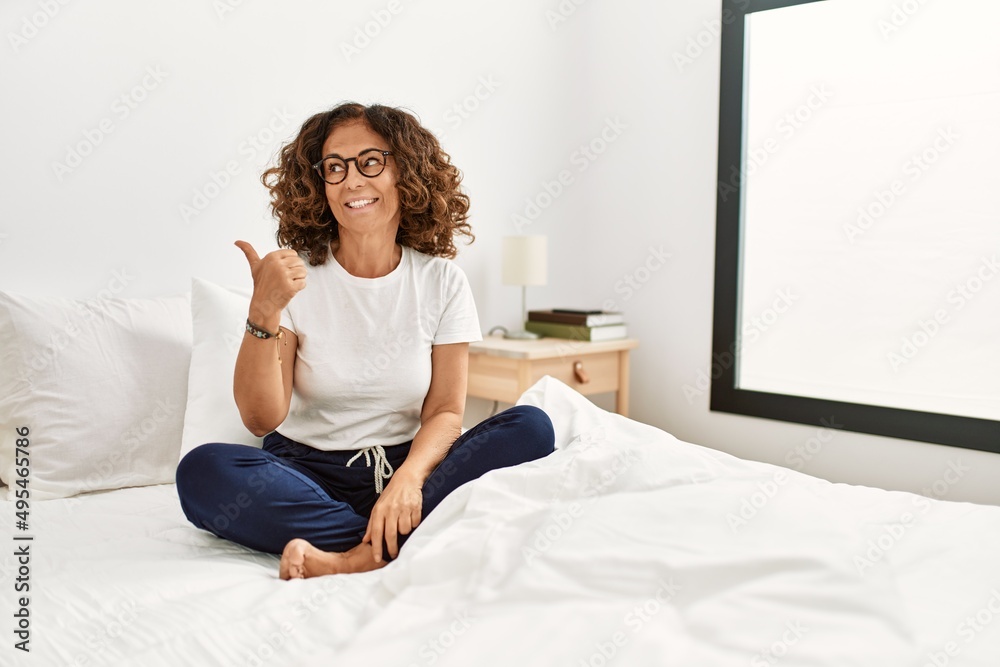 Middle age hispanic woman sitting on the bed at home smiling with happy face looking and pointing to the side with thumb up. Middle age hispanic woman sitting on the bed at home smiling with happy face looking and pointing to the side with thumb up.