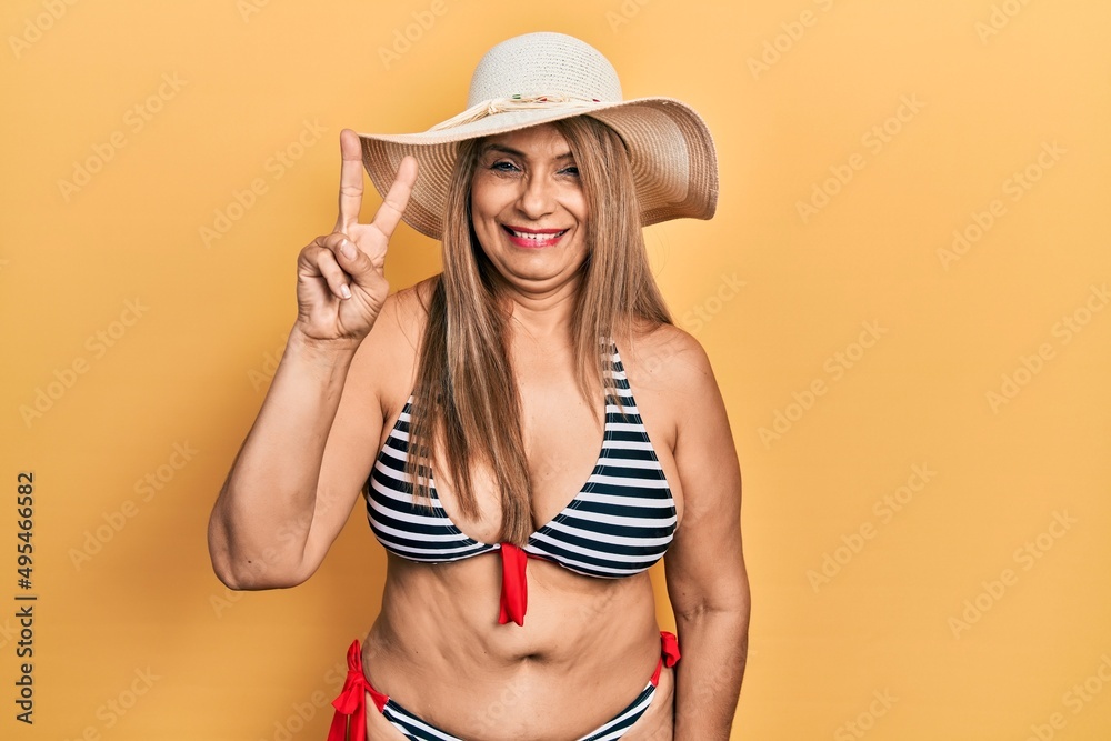 Middle age hispanic woman wearing bikini and summer hat showing and pointing up with fingers number two while smiling confident and happy. Middle age hispanic woman wearing bikini and summer hat showing and pointing up with fingers number two while smiling confident and happy.