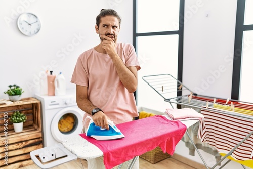 Young hispanic man ironing clothes at home looking stressed and nervous with hands on mouth biting nails. anxiety problem.