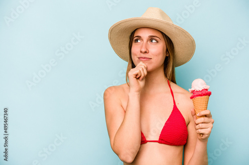 Young caucasian woman wearing a bikini and holding an ice cream isolated on blue background looking sideways with doubtful and skeptical expression.