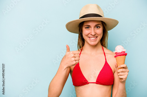 Young caucasian woman wearing a bikini and holding an ice cream isolated on blue background smiling and raising thumb up