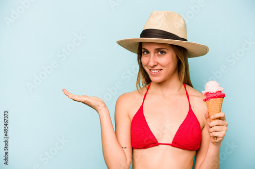 Young caucasian woman wearing a bikini and holding an ice cream isolated on blue background showing a copy space on a palm and holding another hand on waist.
