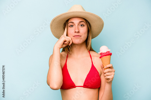 Young caucasian woman wearing a bikini and holding an ice cream isolated on blue background pointing temple with finger, thinking, focused on a task.