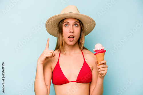 Young caucasian woman wearing a bikini and holding an ice cream isolated on blue background pointing upside with opened mouth.