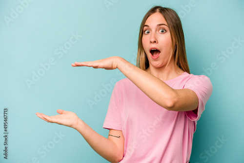 Young caucasian woman isolated on blue background shocked and amazed holding a copy space between hands. © Asier