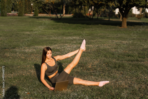 Active fit girl training online with laptop on green grass in the park. Sport lifestyle. Outdoor training.