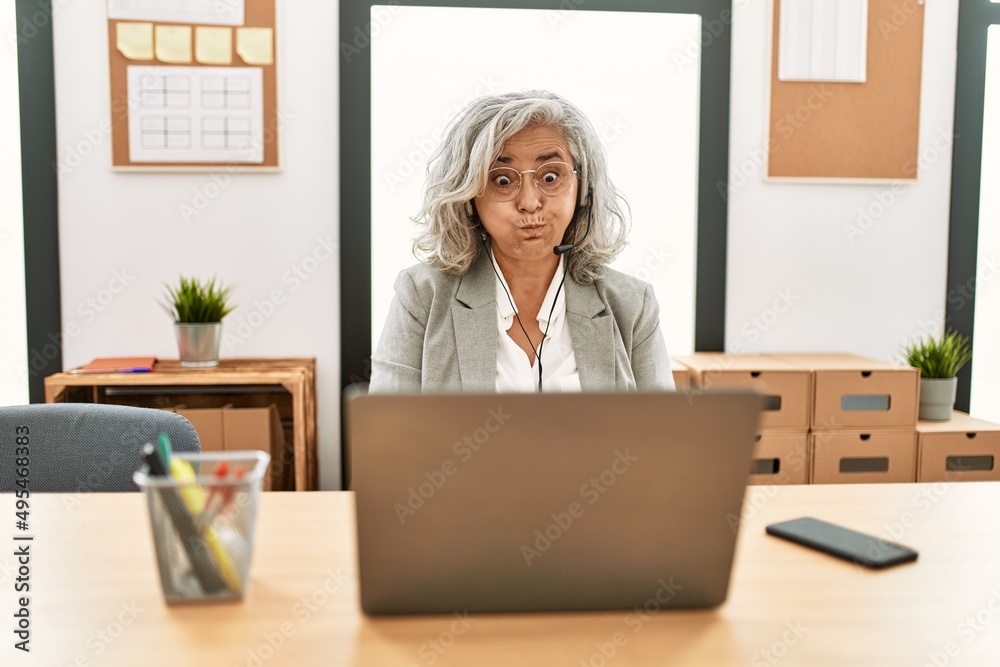 Middle age businesswoman sitting on desk working using laptop at office puffing cheeks with funny face. mouth inflated with air, crazy expression. Middle age businesswoman sitting on desk working using laptop at office puffing cheeks with funny face. mouth inflated with air, crazy expression.