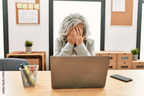 Middle age businesswoman sitting on desk working using laptop at office with sad expression covering face with hands while crying. depression concept.