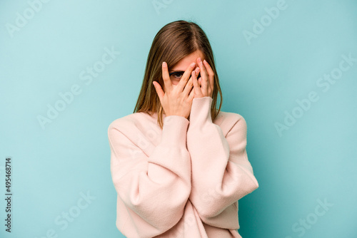 Quadro su tela Young caucasian woman isolated on blue background blink at the camera through fingers, embarrassed covering face