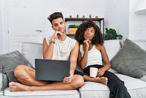 Young interracial couple using laptop at home sitting on the sofa with hand on chin thinking about question, pensive expression. smiling with thoughtful face. doubt concept.