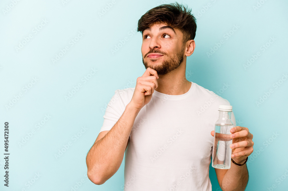 Young hispanic man holding a bottle of water isolated on white background looking sideways with doubtful and skeptical expression.