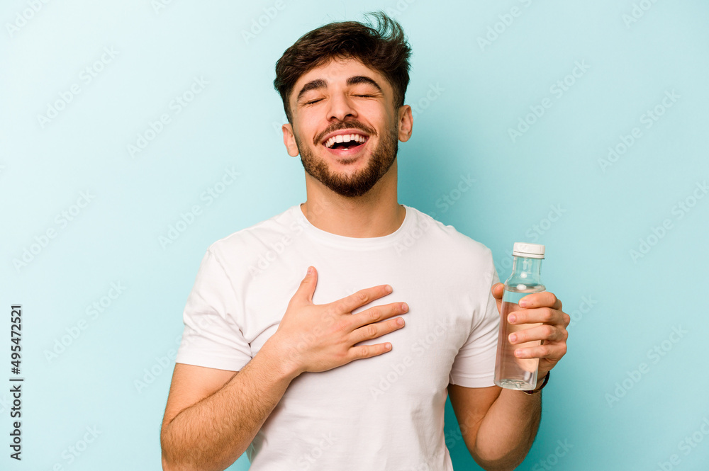 Young hispanic man holding a bottle of water isolated on white background laughs out loudly keeping hand on chest.
