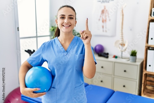 Young physiotherapist woman holding pilates ball at medical clinic surprised with an idea or question pointing finger with happy face  number one
