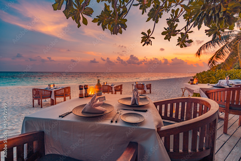Romantic sunset on tropical island shore. Dinner table in beach restaurant. Beautiful sunset sky, tables chairs in sand, close to sea, horizon. Romance, couple honeymoon, wedding destination, vacation