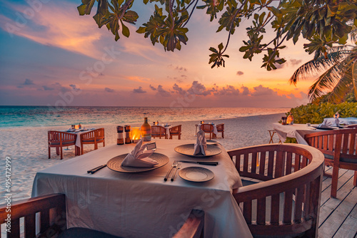 Fototapeta Naklejka Na Ścianę i Meble -  Romantic sunset on tropical island shore. Dinner table in beach restaurant. Beautiful sunset sky, tables chairs in sand, close to sea, horizon. Romance, couple honeymoon, wedding destination, vacation