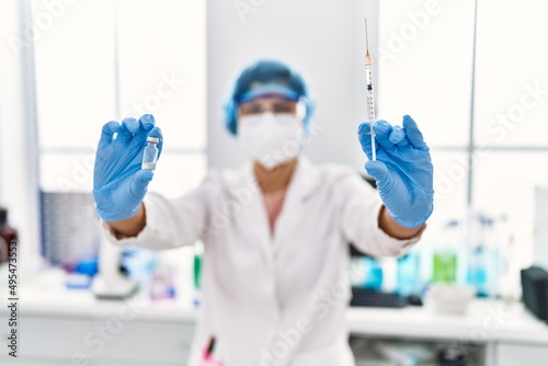 Young latin woman wearing scientist uniform holding vaccine at laboratory
