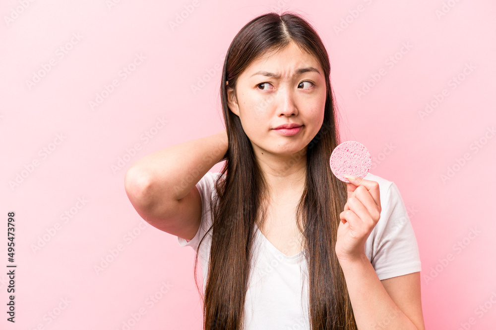 Young asian woman holding facial sponge isolated on pink background touching back of head, thinking and making a choice. Young asian woman holding facial sponge isolated on pink background touching back of head, thinking and making a choice.