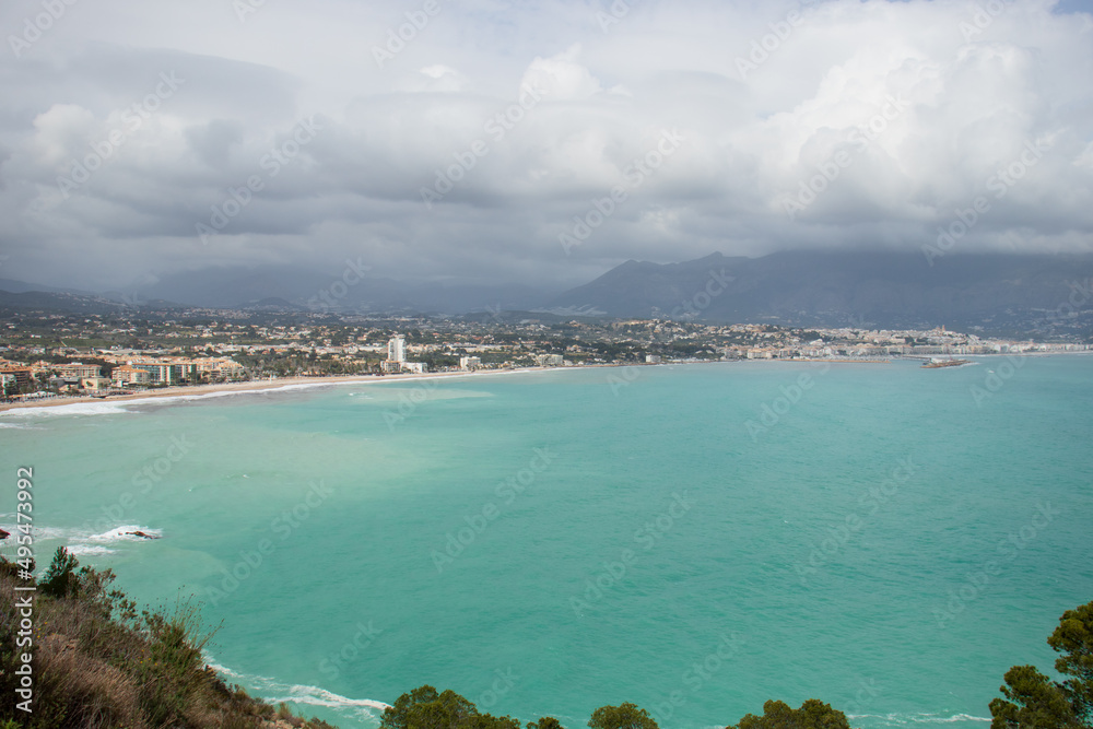 View of the small bay formed by the Racó Albir beach and Cap Blanc beach, in Alicante, during a cloudy day. Turquoise water of the Mediterranean Sea and mountains in the background.