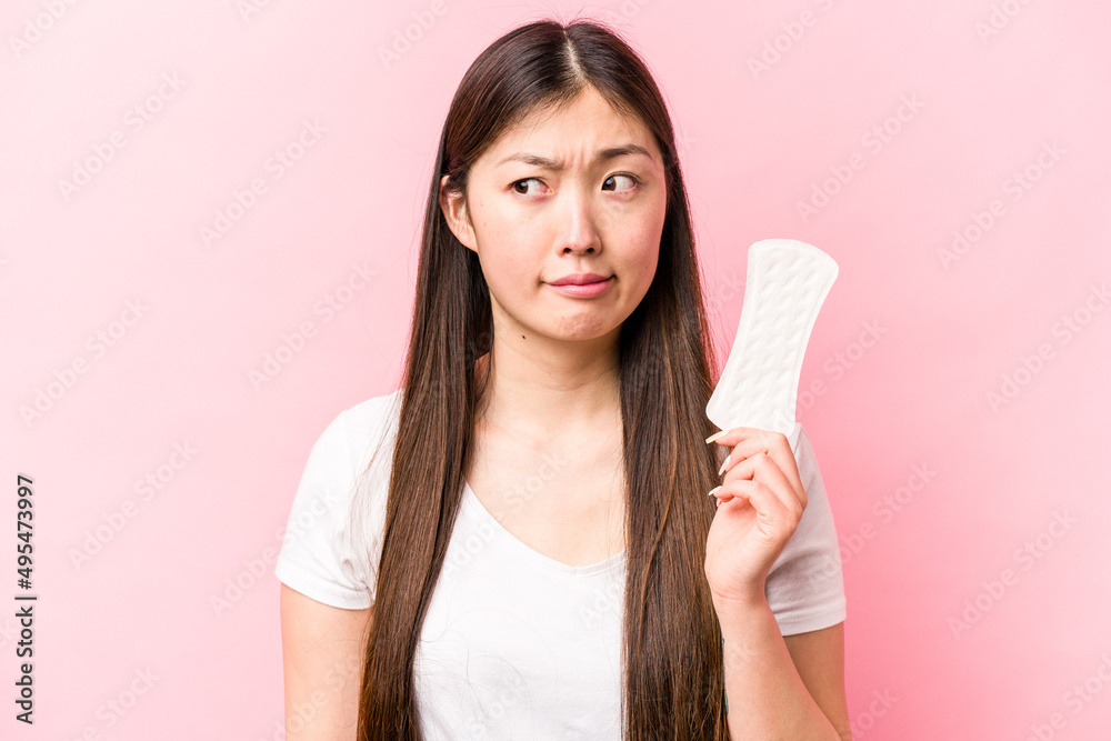 Young asian woman holding sanitary napkin isolated on pink background confused, feels doubtful and unsure. Young asian woman holding sanitary napkin isolated on pink background confused, feels doubtful and unsure.