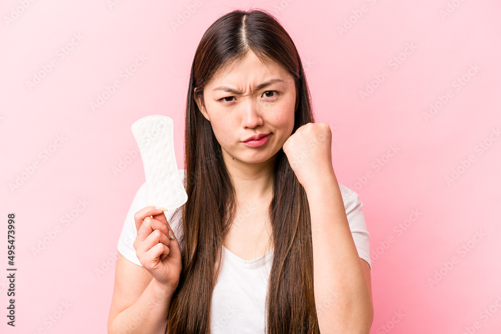 Young asian woman holding sanitary napkin isolated on pink background showing fist to camera, aggressive facial expression. Young asian woman holding sanitary napkin isolated on pink background showing fist to camera, aggressive facial expression.