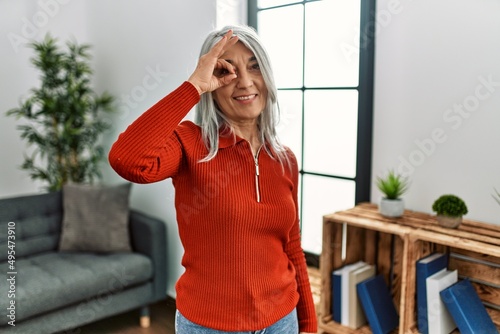 Middle age grey-haired woman wearing casual clothes standing at home smiling happy doing ok sign with hand on eye looking through fingers