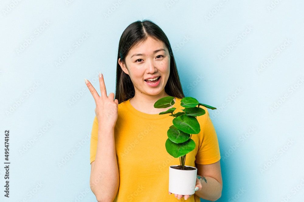 Young asian woman holding a plant isolated on blue background joyful and carefree showing a peace symbol with fingers. Young asian woman holding a plant isolated on blue background joyful and carefree showing a peace symbol with fingers.