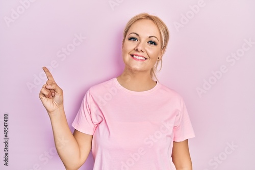 Young blonde woman wearing casual pink t shirt with a big smile on face  pointing with hand finger to the side looking at the camera.