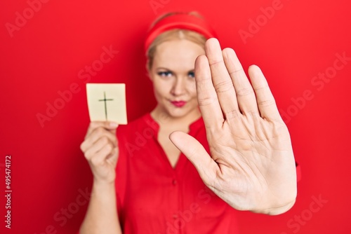 Obraz na plátne Young blonde woman holding catholic cross reminder with open hand doing stop sig