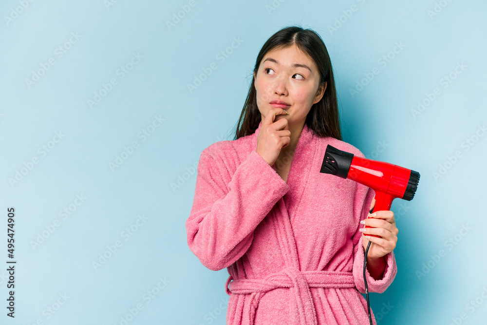 Young asian woman wearing a bathrobe and holding hairdryer isolated on pink background looking sideways with doubtful and skeptical expression. Young asian woman wearing a bathrobe and holding hairdryer isolated on pink background looking sideways with doubtful and skeptical expression.