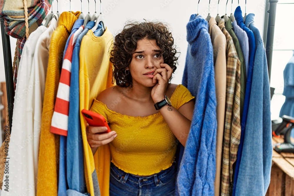 Young hispanic woman searching clothes on clothing rack using smartphone  looking stressed and nervous with hands on mouth biting nails. anxiety  problem. Photos | Adobe Stock