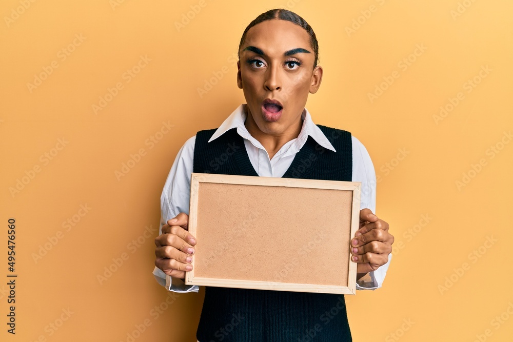 Hispanic man wearing make up and long hair holding empty corkboard afraid and shocked with surprise and amazed expression, fear and excited face. Hispanic man wearing make up and long hair holding empty corkboard afraid and shocked with surprise and amazed expression, fear and excited face.