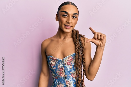 Hispanic man wearing make up and long hair wearing elegant corset smiling and confident gesturing with hand doing small size sign with fingers looking and the camera. measure concept.