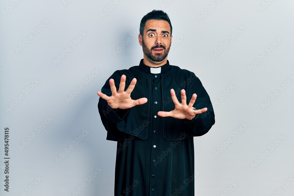 Handsome hispanic man with beard wearing catholic priest robe afraid and terrified with fear expression stop gesture with hands, shouting in shock. panic concept. Handsome hispanic man with beard wearing catholic priest robe afraid and terrified with fear expression stop gesture with hands, shouting in shock. panic concept.