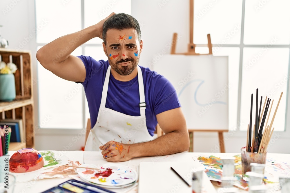 Young hispanic man with beard at art studio with painted face confuse and wonder about question. uncertain with doubt, thinking with hand on head. pensive concept.