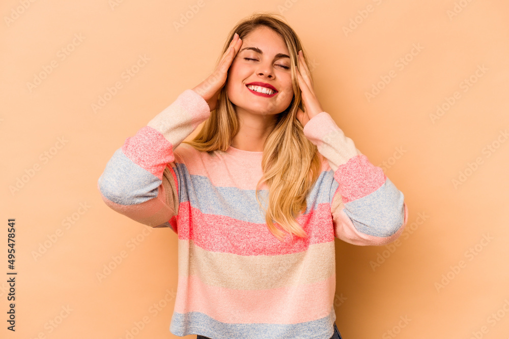 Young caucasian woman isolated on beige background laughs joyfully keeping hands on head. Happiness concept. Young caucasian woman isolated on beige background laughs joyfully keeping hands on head. Happiness concept.