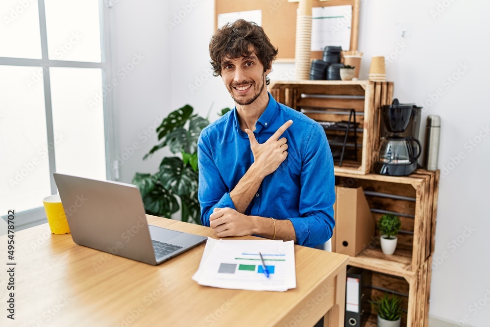 Young hispanic man with beard working at the office using computer laptop cheerful with a smile of face pointing with hand and finger up to the side with happy and natural expression on face