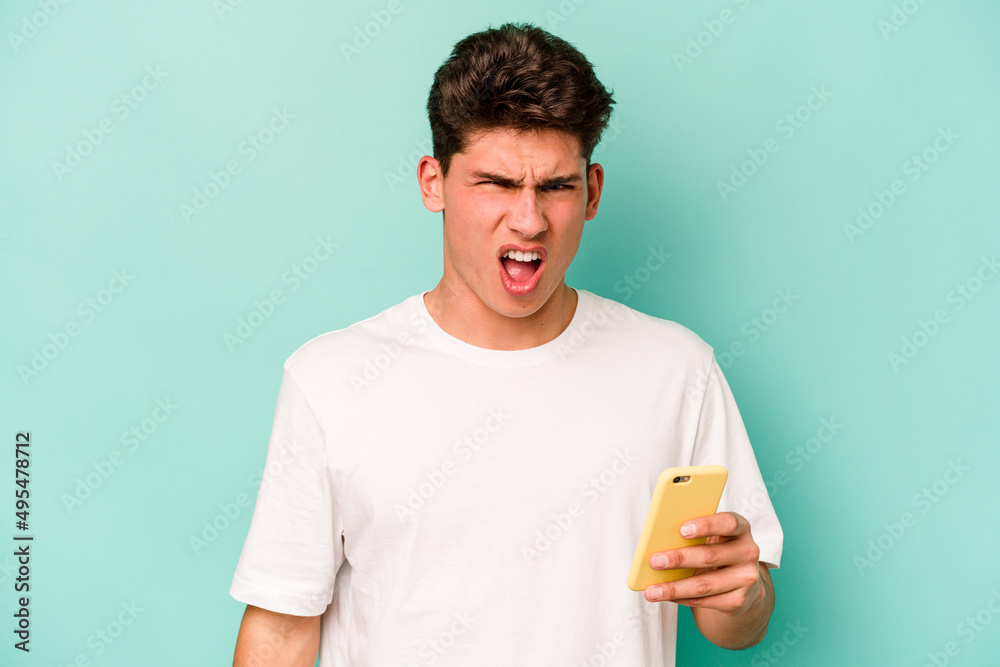 Young caucasian man holding mobile phone isolated on blue background screaming very angry and aggressive. Young caucasian man holding mobile phone isolated on blue background screaming very angry and aggressive.