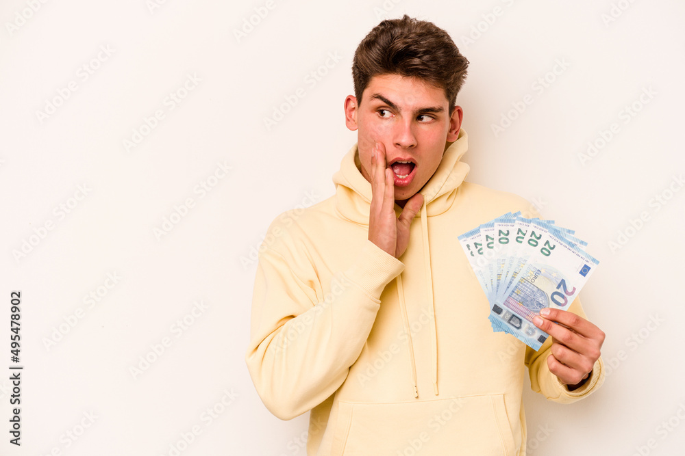 Young caucasian man holding banknotes isolated on white background is saying a secret hot braking news and looking aside Young caucasian man holding banknotes isolated on white background is saying a secret hot braking news and looking aside