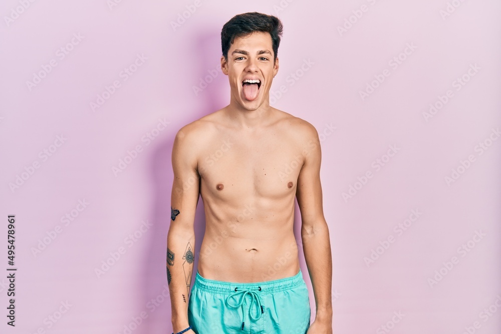 Young hispanic man wearing swimwear shirtless sticking tongue out happy with funny expression. emotion concept.